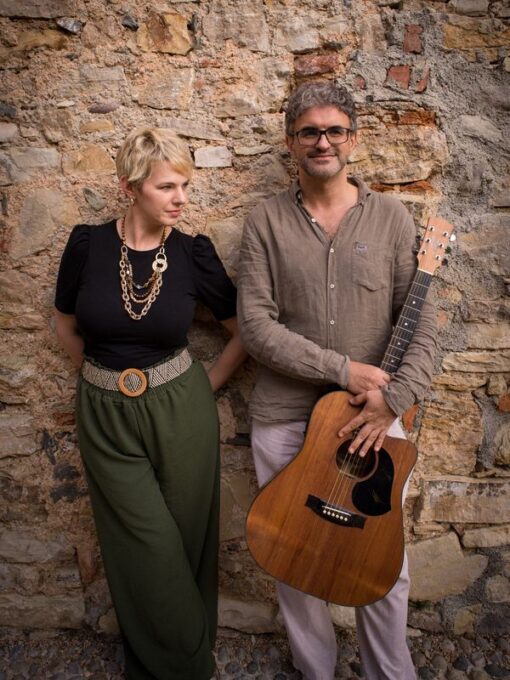 Un uomo e una donna con chitarre accanto a un muro di pietra.